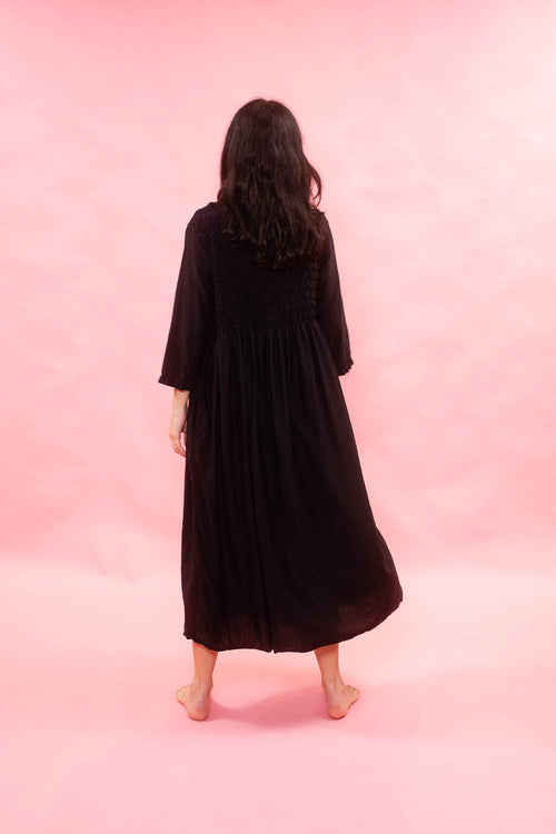 A woman viewed from behind wearing a long black midi dress with three-quarter sleeves, standing barefoot against a solid pink studio backdrop.