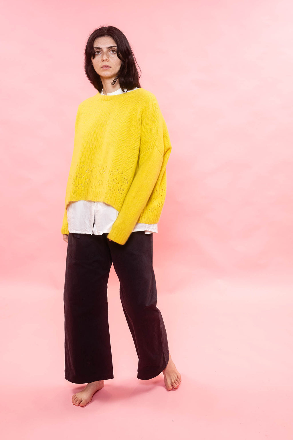 Person wearing a bright yellow cashmere oversized hand knit 
sweater over a white shirt with dark pants on a pink background
