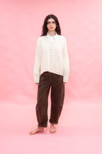 A woman stands barefoot against a solid pink studio backdrop, wearing a cream button-front cashmere hand knit oversized cardigan over a white collared shirt and dark brown corduroy pants.

