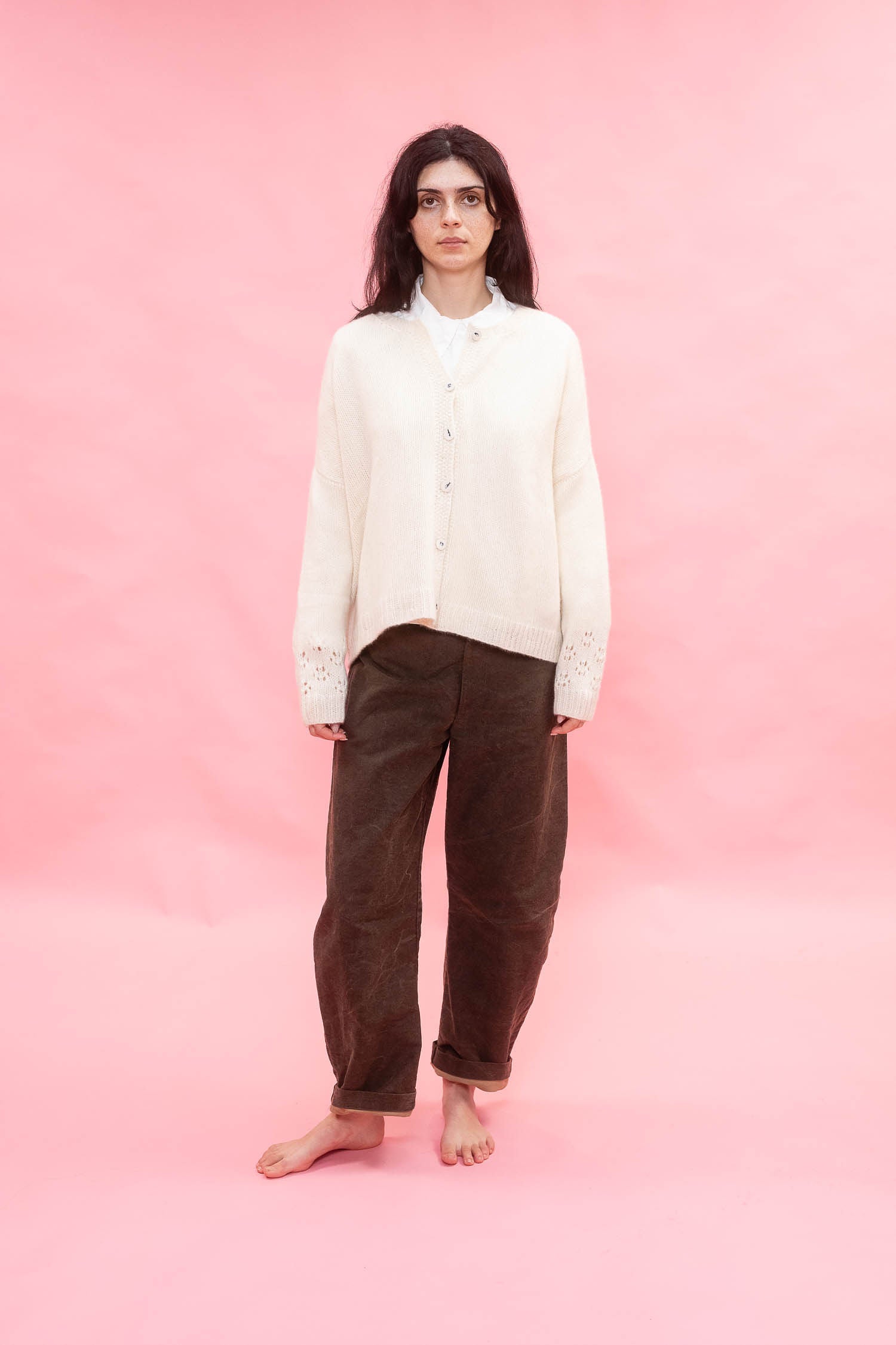 A woman stands barefoot against a solid pink studio backdrop, wearing a cream button-front cashmere hand knit oversized cardigan over a white collared shirt and dark brown corduroy pants.
