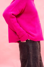 A close-up side view of a person wearing a bright neon pink hand knit cashmere cardigan sweater with dark charcoal pants, set against a solid light pink background