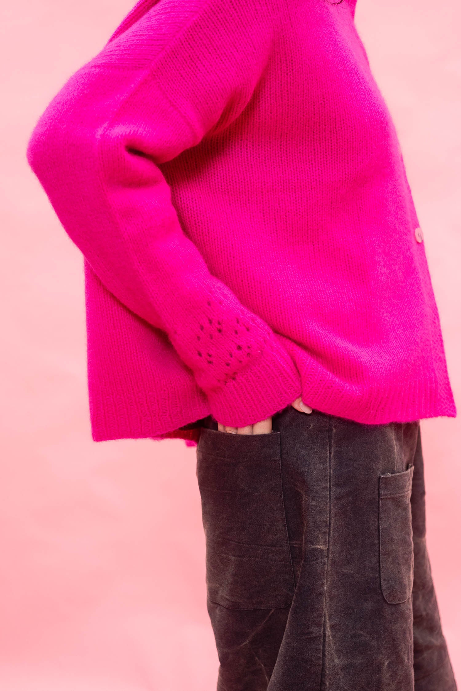 A close-up side view of a person wearing a bright neon pink hand knit cashmere cardigan sweater with dark charcoal pants, set against a solid light pink background
