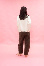 A person standing with their back to the camera, wearing a cream-colored hand knit cashmere oversized cardigan sweater and dark brown  pants, set against a solid pink studio backdrop.
