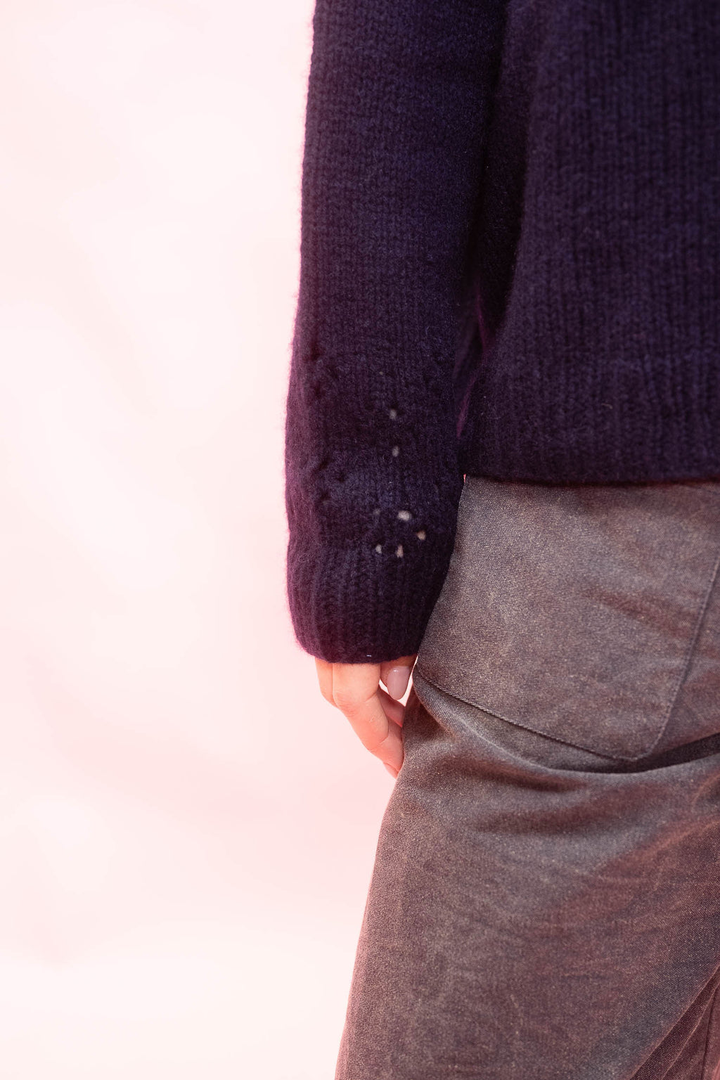 Close-up of a person wearing a dark navy cashmere hand knitted cardigan oversized sweater and grey pants, with one hand in a pocket against a soft pink background.