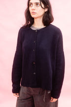 A woman wearing a dark navy blue knit button-up hand knitted cashmere oversized cardigan sweater over a grey shirt, paired with brown pants, set against a solid pink background.
