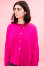 A woman wearing a bright fuchsia neon pink  hand knit cashmere cardigan with a button-up front, photographed against a solid light pink background.
