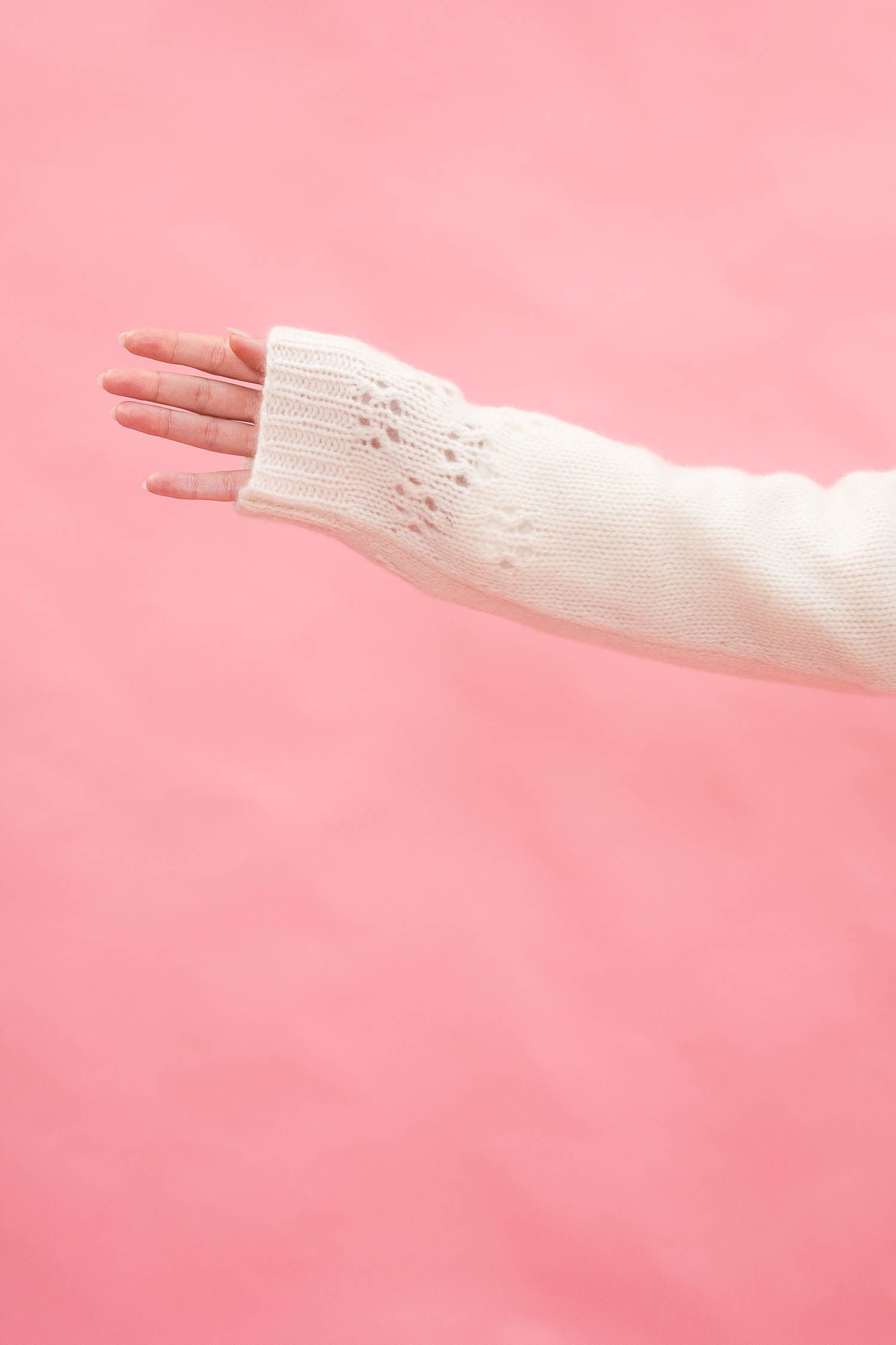 A close-up of a person's hand and forearm wearing a cream-coloured hand knit sleeve with delicate lace detailing, in cashmere, set against a solid pink background.
