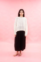 A model stands barefoot against a solid pink studio background, wearing an oversized cream handknit cashmere sweater with a decorative lace stitch at the hem and a long dark skirt.