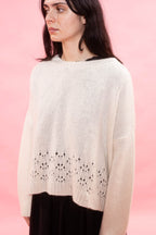 A woman wearing a cream hand knit cashmere oversized sweater with a lace stitch along the hem, set against a solid pink background.

