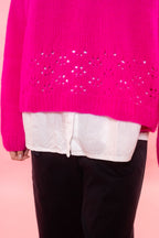 A close-up of a woman wearing a bright hot pink knit sweater with decorative knit details across the lower part, layered over a white top and black pants, against a solid pink background.