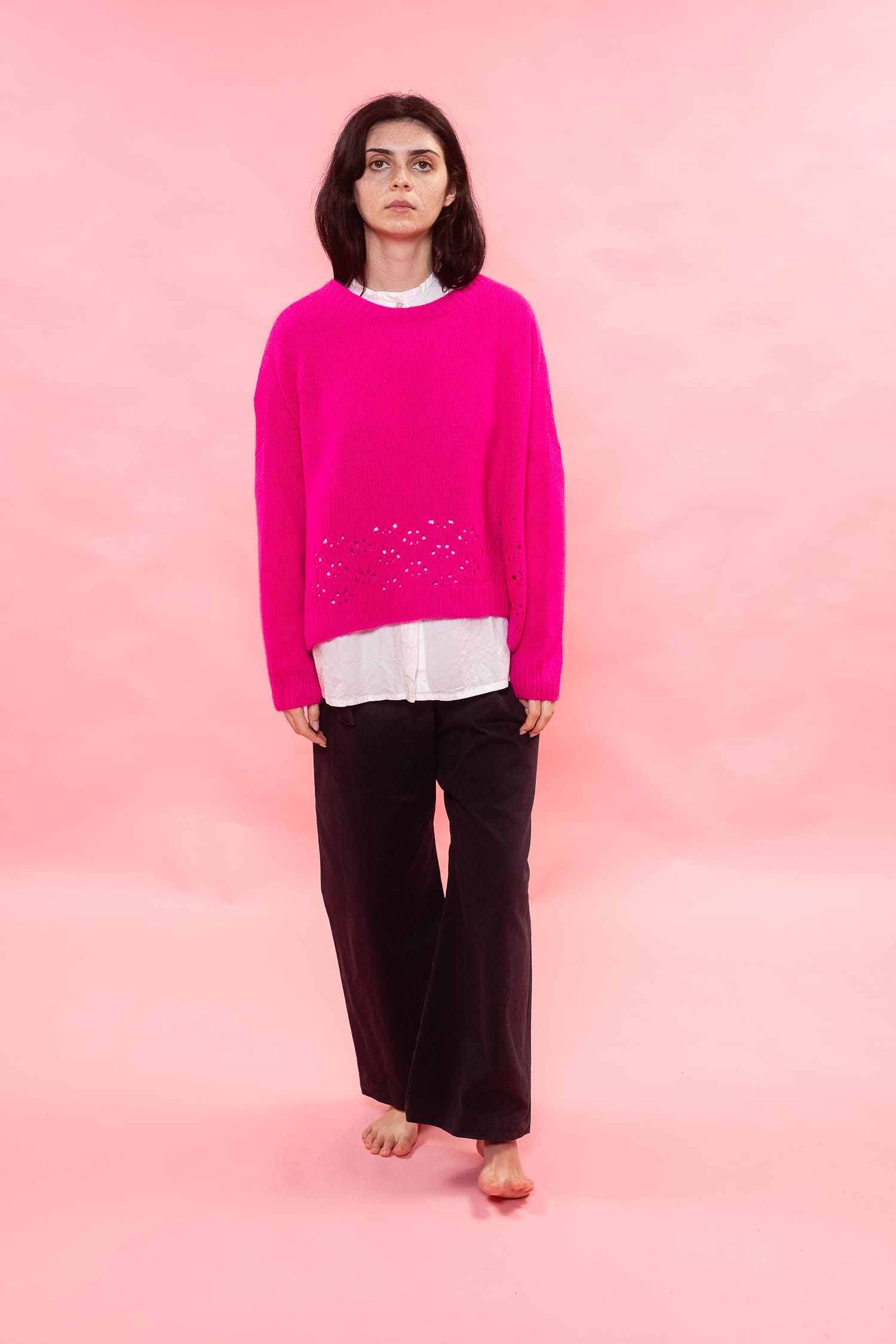 A woman stands against a solid pink studio background wearing a bright fuchsia pink hand knit cashmere sweater with a lace stitch pattern across the lower front, layered over a white shirt and dark brown wide-leg trousers