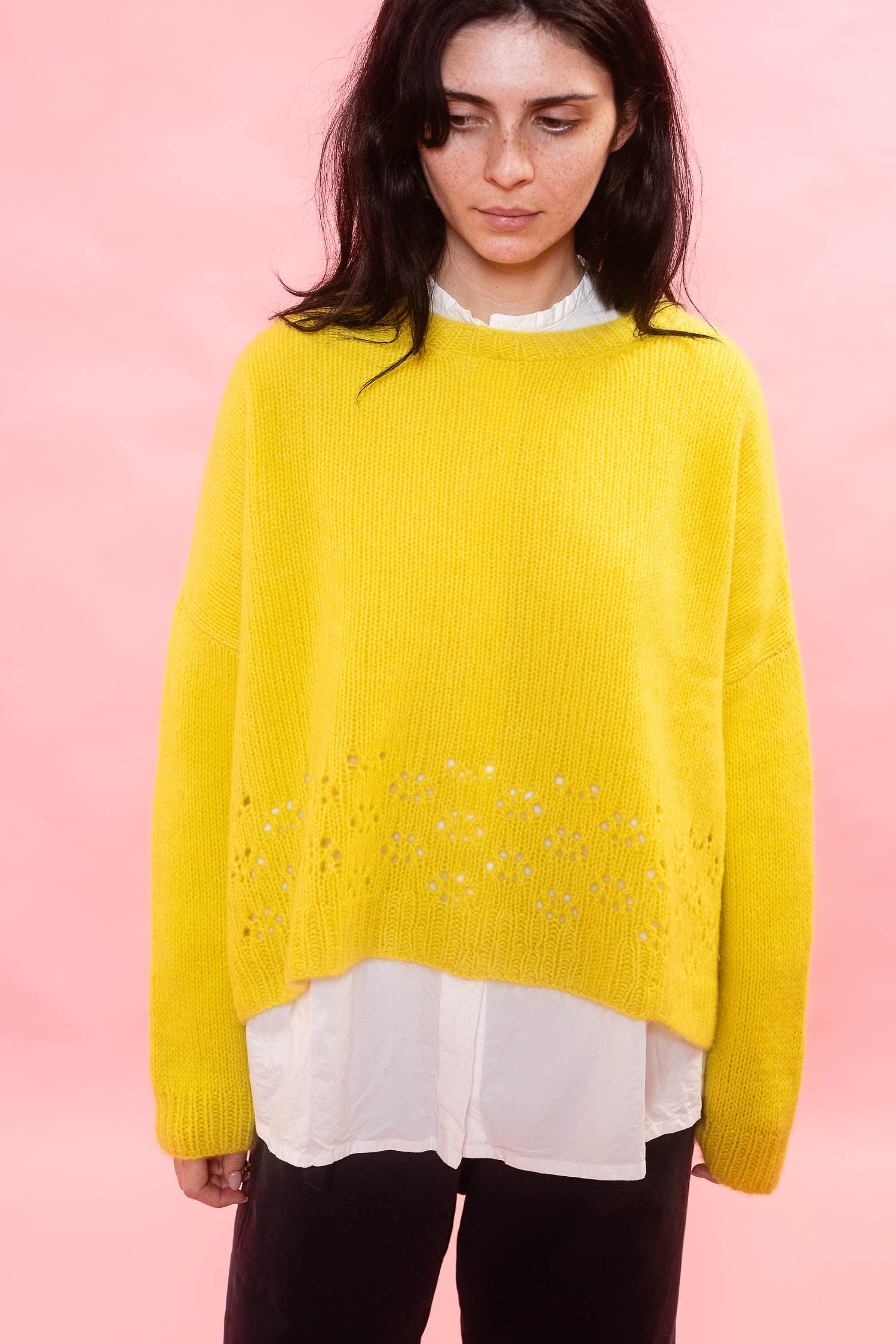 A woman wearing a bright yellow oversized hand knit cashmere sweater with a lace stitch pattern across the lower half, layered over a white collared shirt and black pants, set against a solid pink background.