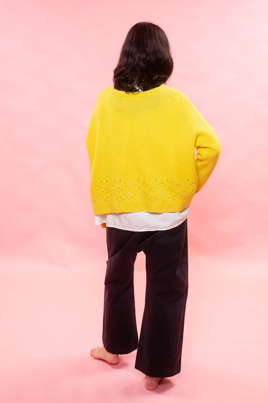 A person shown from the back wearing a bright lemon yellow hand knit cashmere sweater with a lace knit hem, layered over a white shirt and dark brown wide-leg pants, standing against a solid pink background.

