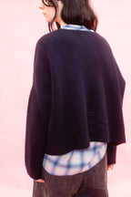 A person shown from the back wearing a dark navy hand knit cashmere sweater over a blue and white plaid shirt, set against a solid light pink background.