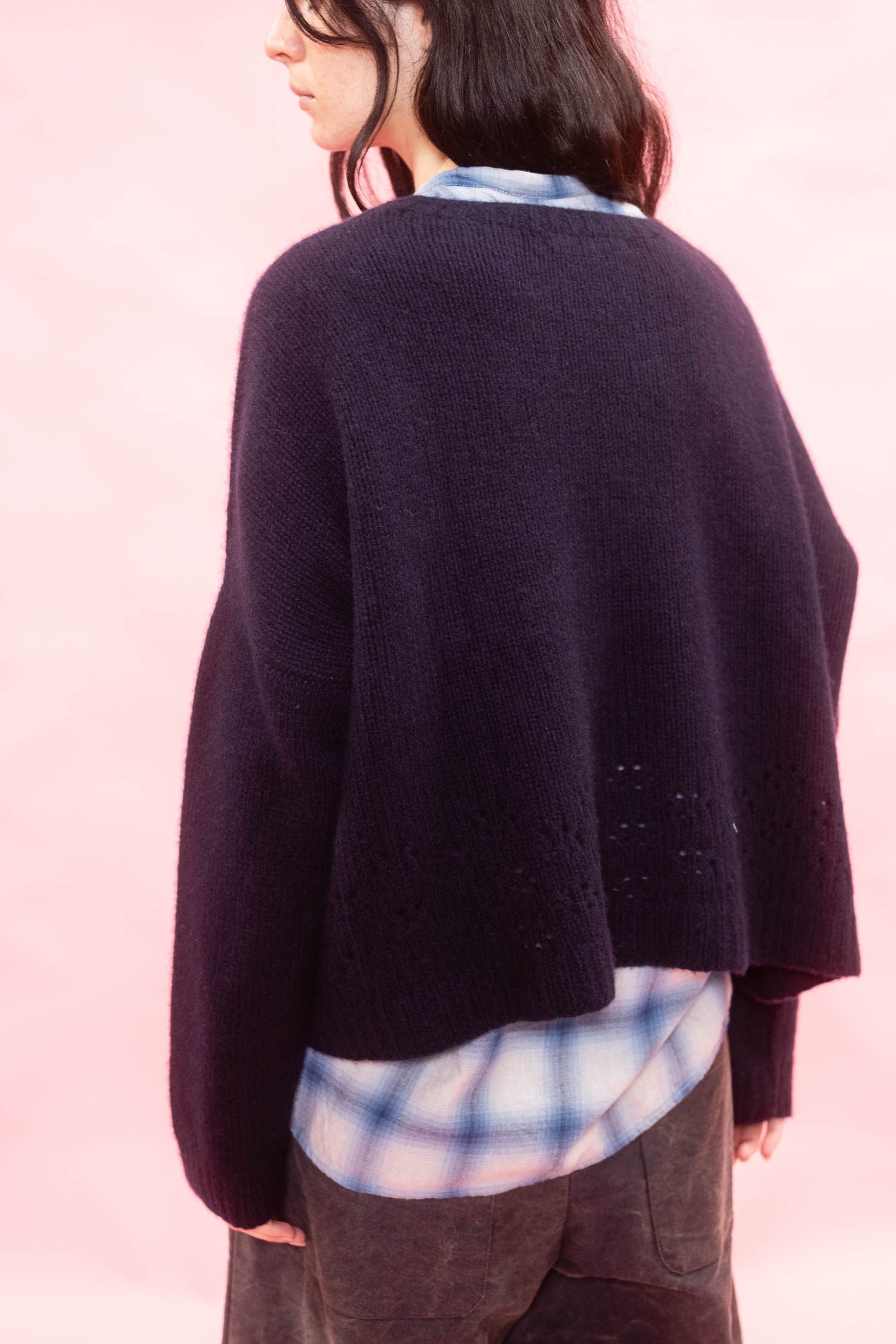 A person shown from the back wearing a dark navy hand knit cashmere sweater over a blue and white plaid shirt, set against a solid light pink background.