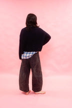 A person standing with their back to the camera, wearing a dark navy  hand knitted cashmere swearer over a blue and white plaid shirt tucked in, and dark wide-leg 
pants against a solid pink background.