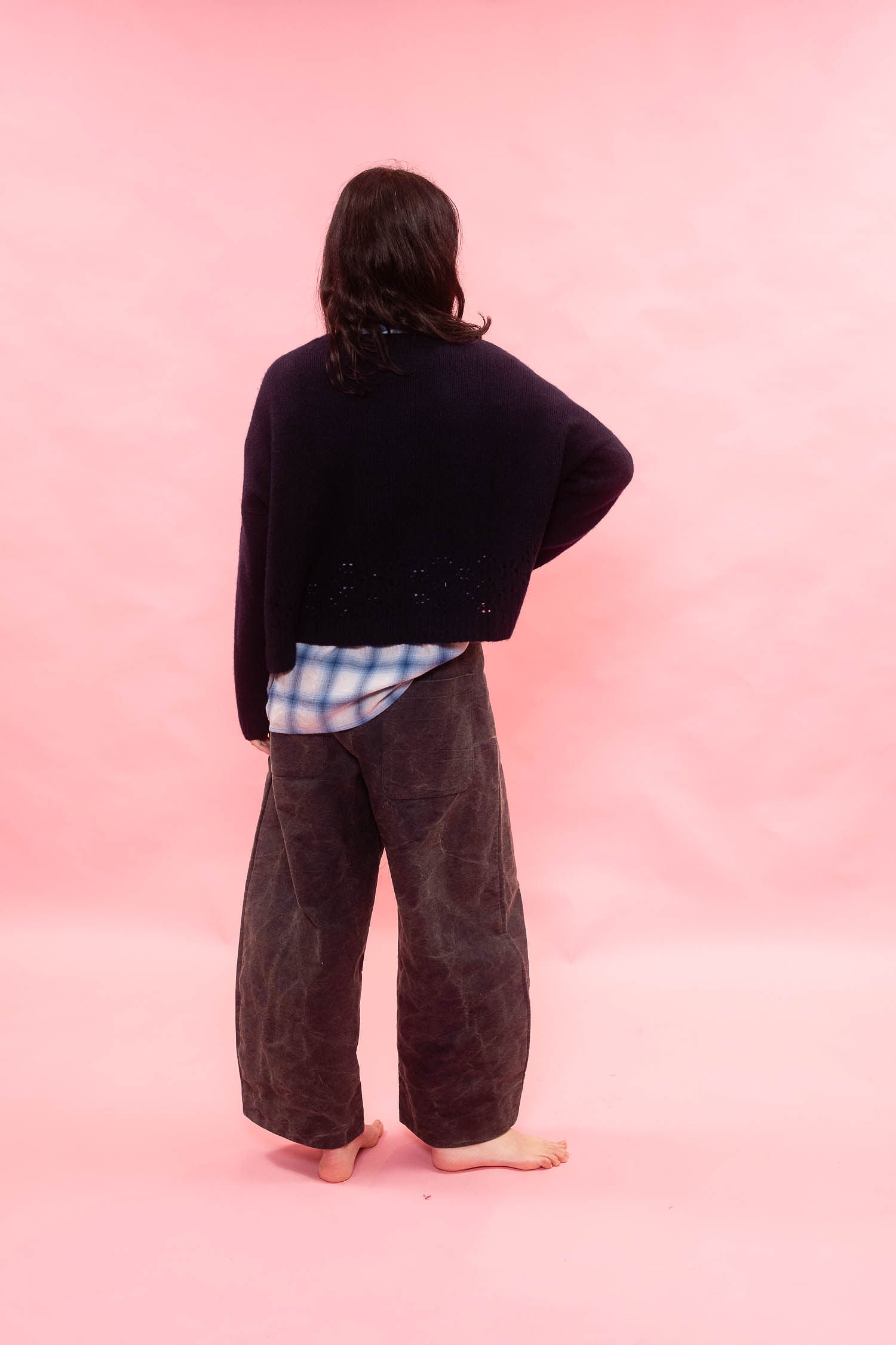 A person standing with their back to the camera, wearing a dark navy  hand knitted cashmere swearer over a blue and white plaid shirt tucked in, and dark wide-leg 
pants against a solid pink background.