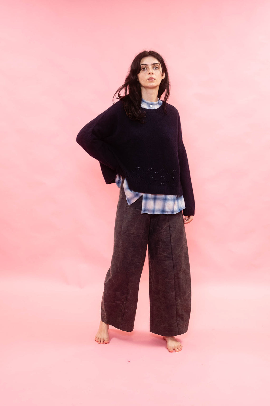 A woman stands barefoot against a solid pink studio backdrop, wearing a dark oversized knit cashmere handknit sweater over a blue-and-white plaid shirt and dark wide-leg trousers.