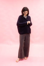 A woman stands barefoot against a solid pink studio backdrop, wearing a dark navy button-front cashmere hand knitted oversized cardigan over a light gray top and dark charcoal wide-leg pants.
