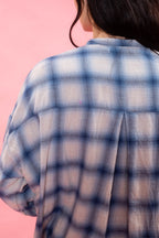 A close-up back view of a person wearing a loose blue and white plaid button-up shirt made in soft Japanese brushed cotton wool mix, standing against a solid pink background.
