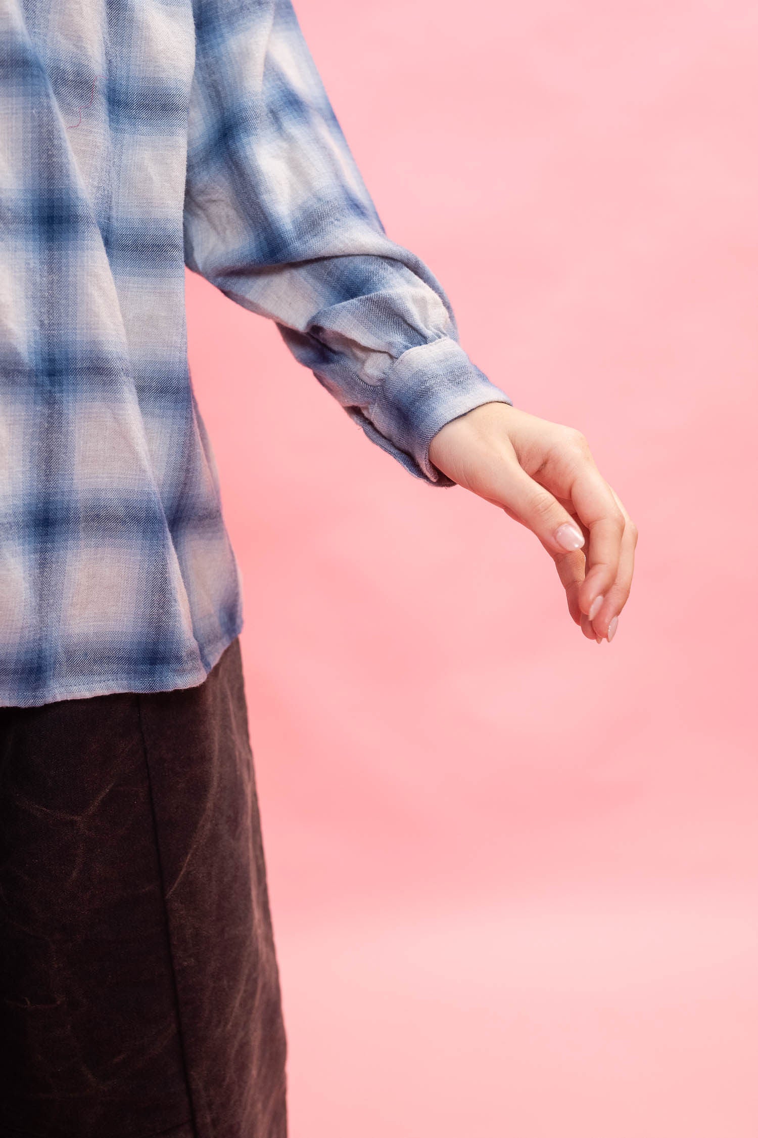 A person wearing a blue and white plaid long-sleeve shirt made from Japanese brushed cotton wool mix fabric, and a dark brown pants shown from the chest down against a solid pink background.
