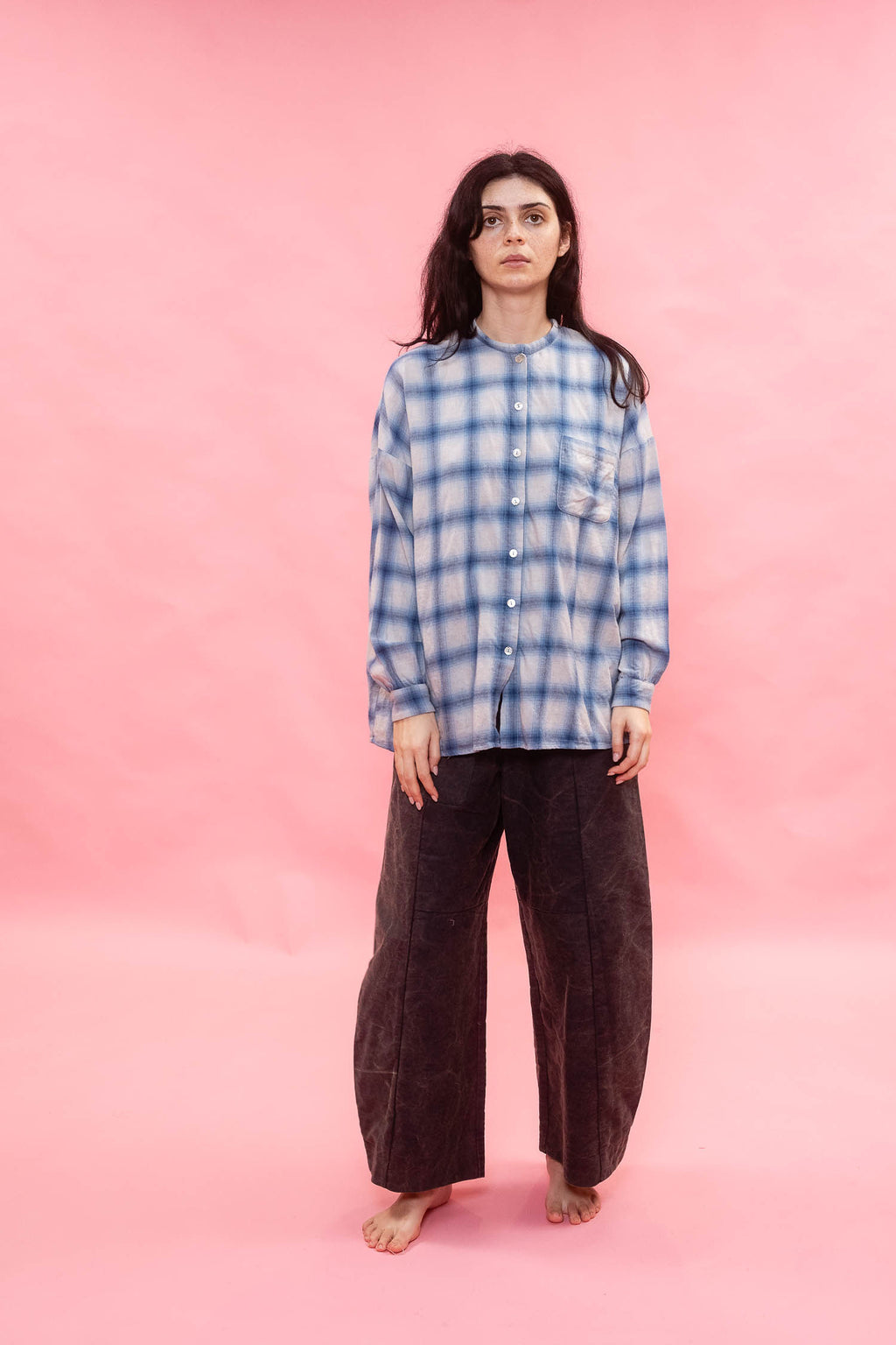 A woman stands barefoot against a solid pink studio backdrop, wearing a blue and white plaid button-up shirt made from Japanese brushed cotton wool mix fabric, with dark brown wide-leg pants.
