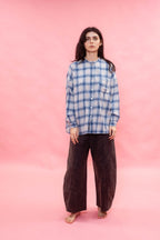 A woman stands barefoot against a solid pink studio backdrop, wearing a blue and white plaid button-up shirt made from Japanese brushed cotton wool mix fabric, with dark brown wide-leg pants.
