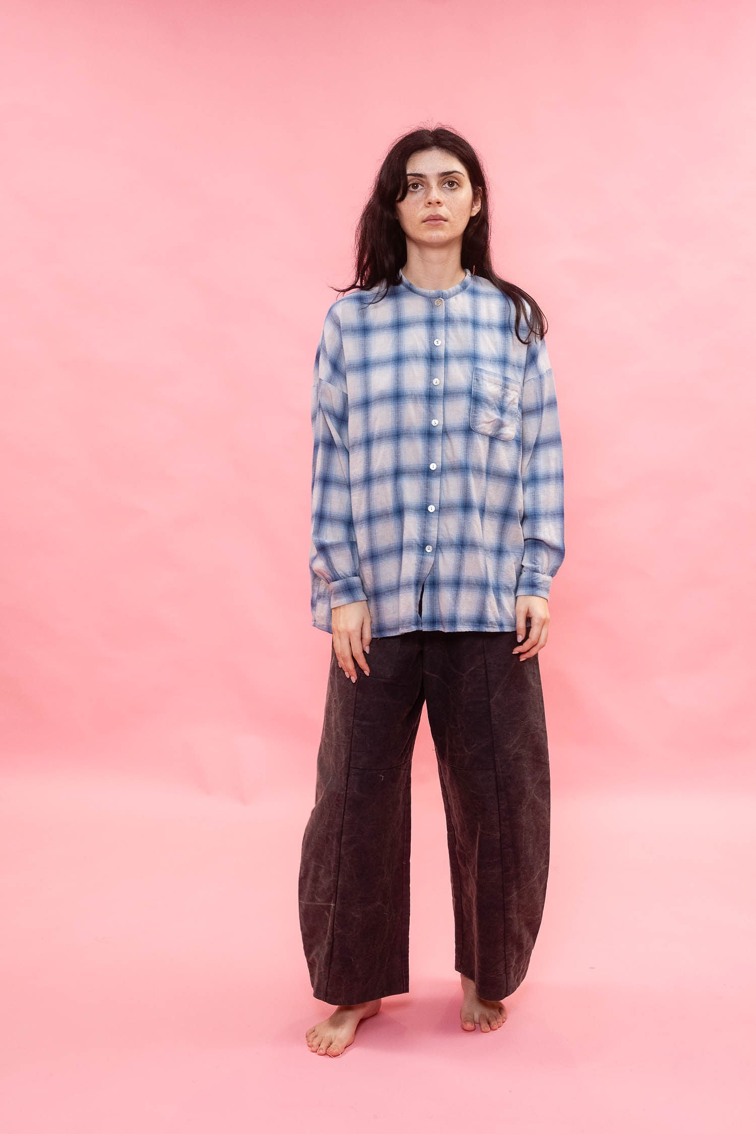 A woman stands barefoot against a solid pink studio backdrop, wearing a blue and white plaid button-up shirt made from Japanese brushed cotton wool mix fabric, with dark brown wide-leg pants.
