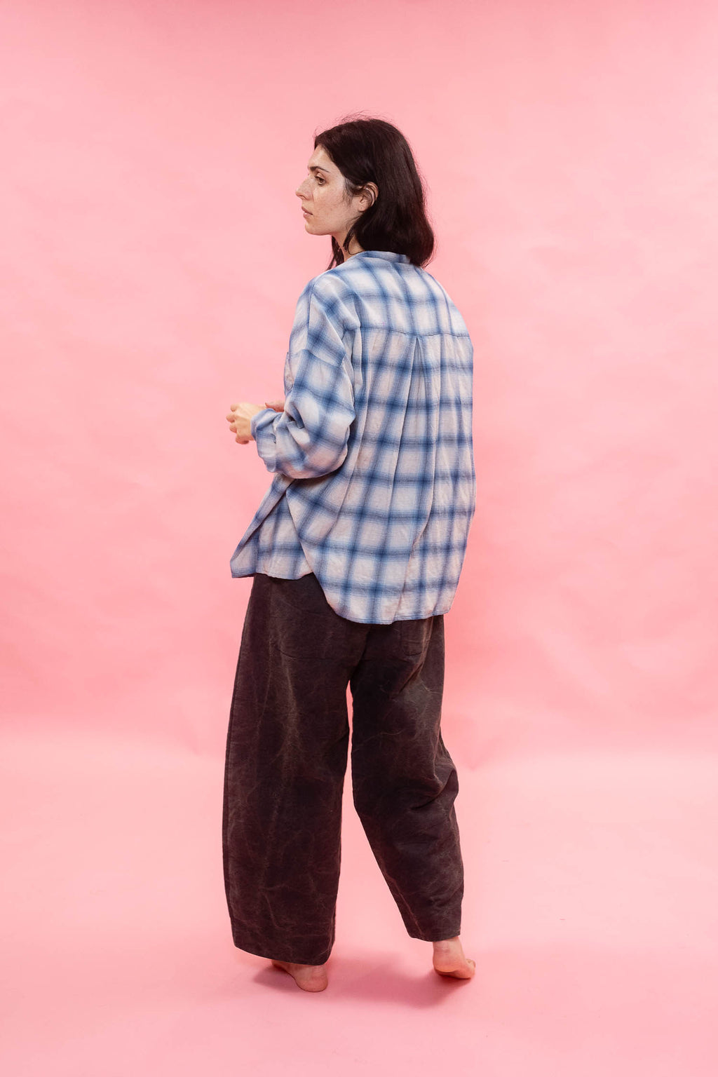 A woman stands barefoot against a solid pink backdrop, wearing a blue and white plaid button-up shirt made from Japanese brushed cotton wool fabric and loose dark brown pants, viewed from the back.
