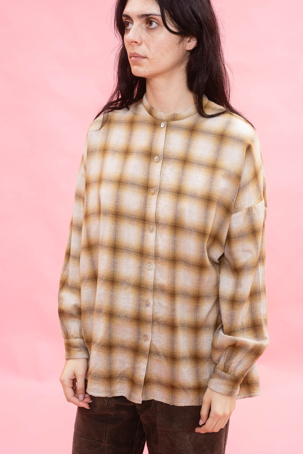 A woman wearing a long-sleeve button-up shirt with a brown and ochre plaid pattern made from Japanese brushed cotton wool fabric, posed against a solid pink background.
