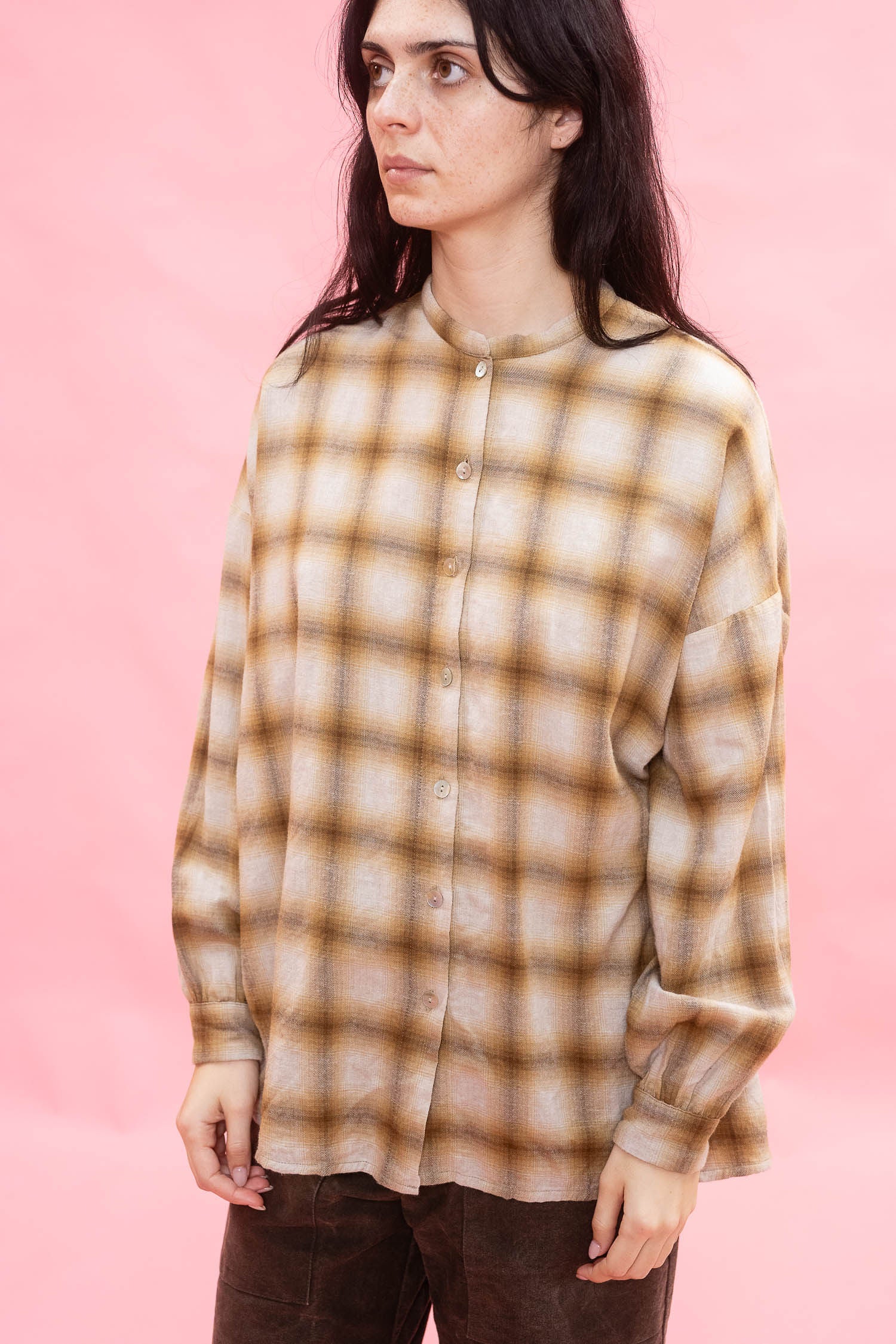 A woman wearing a long-sleeve button-up shirt with a brown and ochre plaid pattern made from Japanese brushed cotton wool fabric, posed against a solid pink background.
