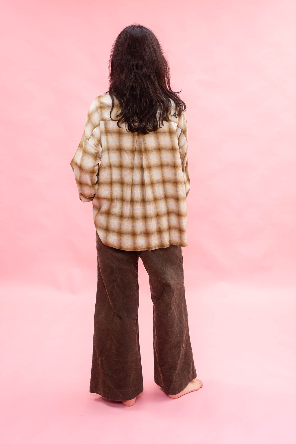 A person standing with their back to the camera, wearing an oversized tan and ochre plaid brushed cotton/wool shirt and dark brown wide-leg pants, set against a solid pink studio background.

