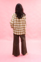 A person standing with their back to the camera, wearing an oversized tan and ochre plaid brushed cotton/wool shirt and dark brown wide-leg pants, set against a solid pink studio background.
