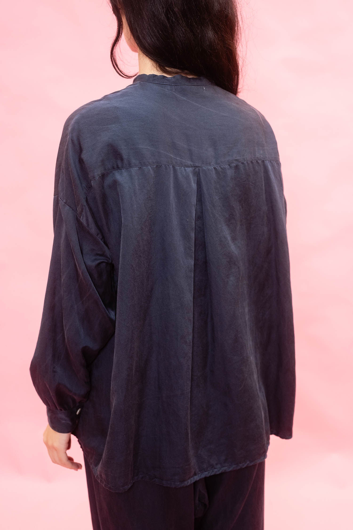 A person shown from the back wearing a loose, dark navy long-sleeve shirt against a solid pink background. made in silk cotton satin. 
