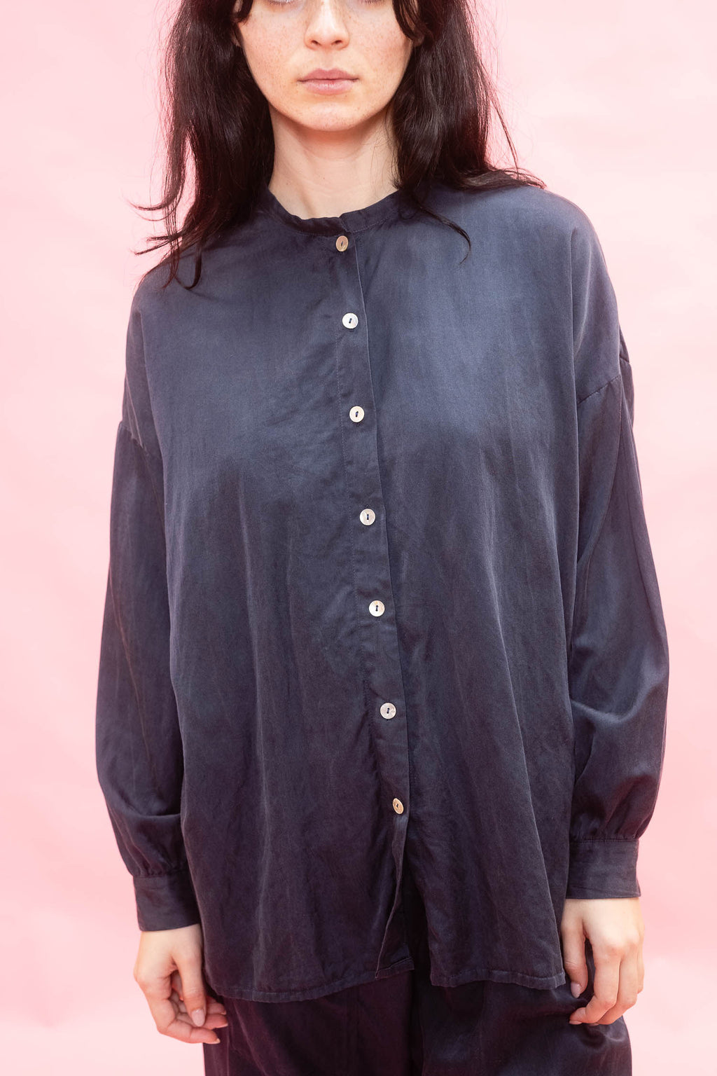 A woman wearing a loose, dark navy long-sleeve button-up shirt with a high collar, photographed against a solid pink background. made in silk cotton satin. 