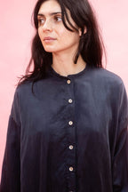A woman with dark hair wearing a dark navy button-up shirt, in silk cotton satin 
shown from the chest up against a solid pink background.
