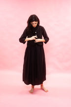 Woman wearing a black dress with a black softly striped wool linen waistcoat. standing against a pink background. 