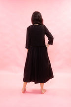 A woman is shown from behind wearing a black long-sleeve midi dress with a black chalk striped waistcoat, standing against a solid pink studio backdrop.