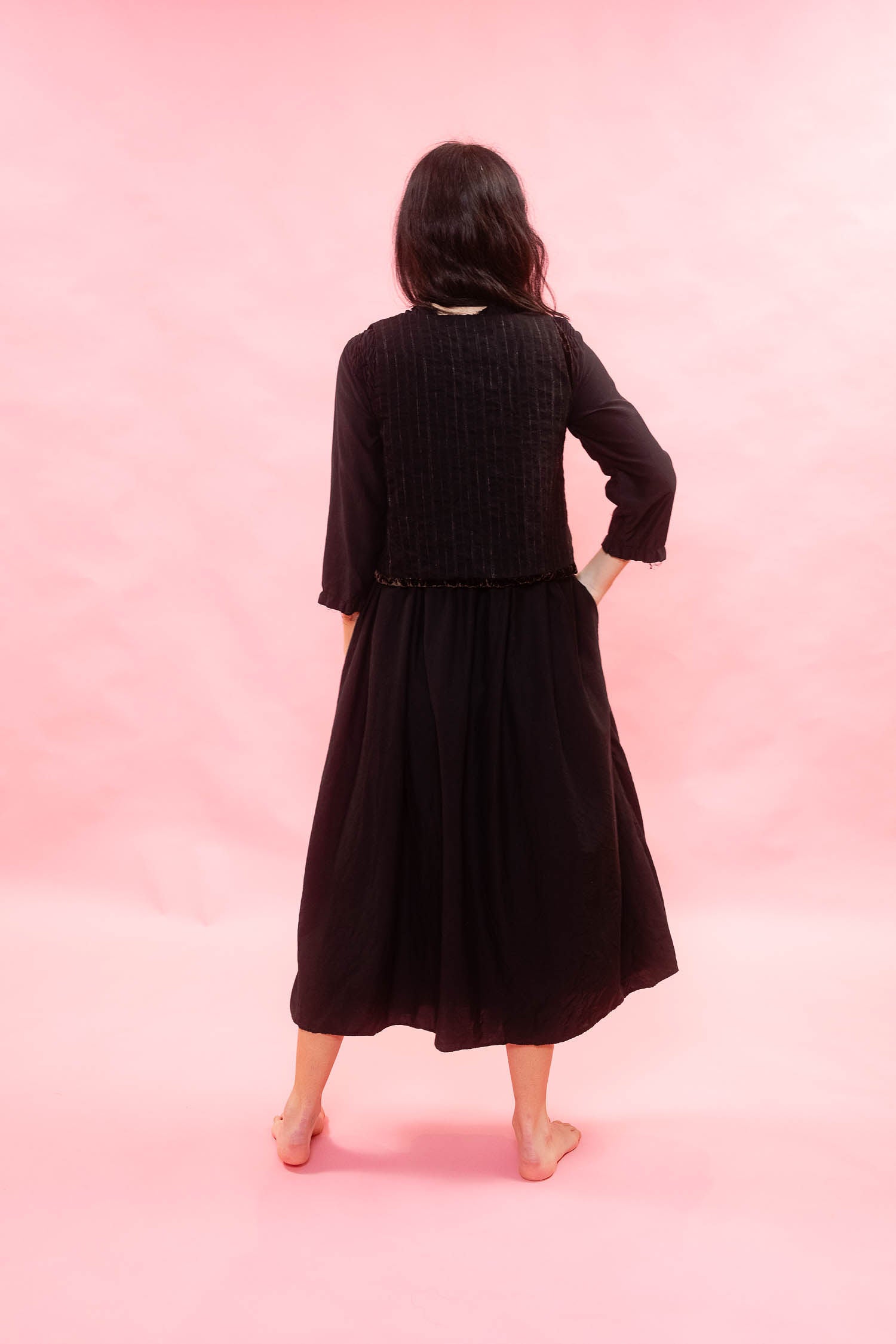 A woman is shown from behind wearing a black long-sleeve midi dress with a black chalk striped waistcoat, standing against a solid pink studio backdrop.
