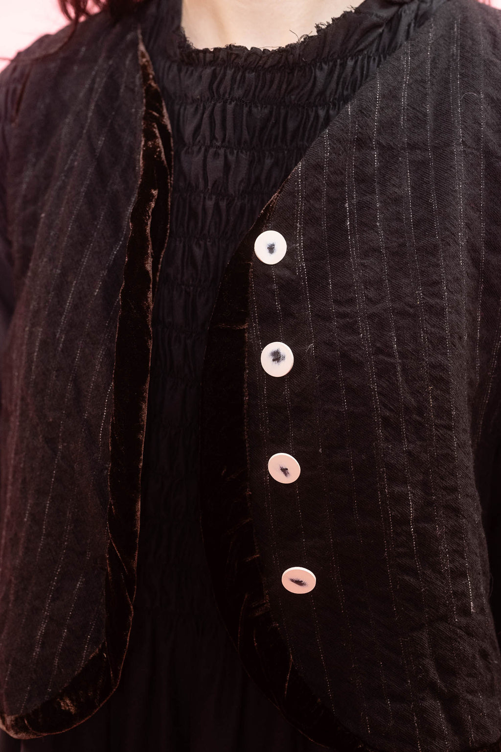 Close-up of a black textured waistcoat  with vertical pinstripes and four light-colored buttons, worn over a matching black smocked top.