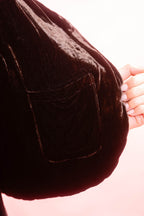 Close-up of a dark brown velvet waistcoat with a pocket, held by a hand with manicured nails against a soft pink background.