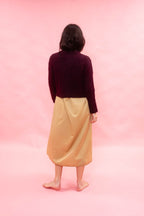 A person standing with their back to the camera, wearing a dark plumknit cropped alpaca cardiga nand a shiny mustard yellow dress, set against a solid pink studio background.
