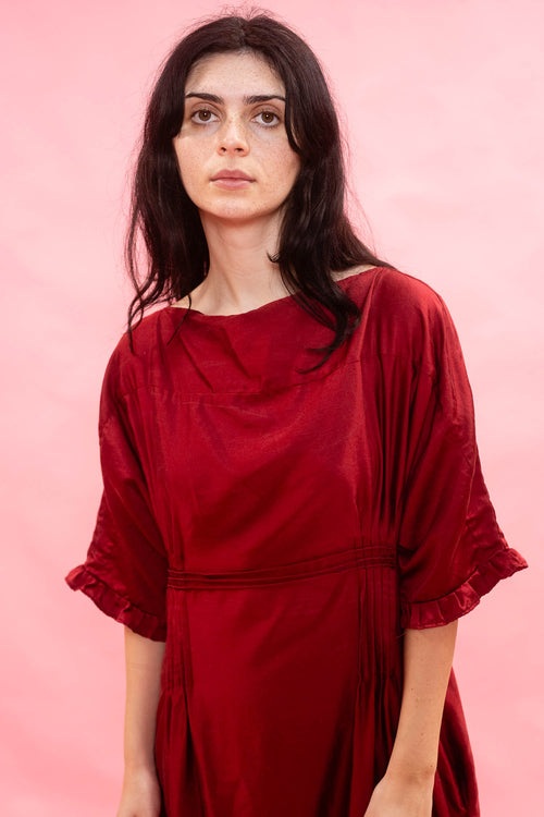 A woman with dark hair wearing a deep red, loose-fitting dress with three-quarter sleeves with a frill on the cuff, standing against a solid pink background.