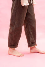 A woman wearing loose dark dirt green brown cotton pants with rolled cuffs, shown from the waist down against a solid pink background.
