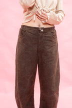 A woman wearing dark brown cotton patina, elasticated waist tapered-leg pants with a wooden button fastening and a cream striped long-sleeve jersey top, standing against a solid pink background.