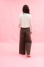 A woman viewed from behind wearing a cream long-sleeve top and dark brown wide-leg pants, standing against a solid pink studio backdrop.