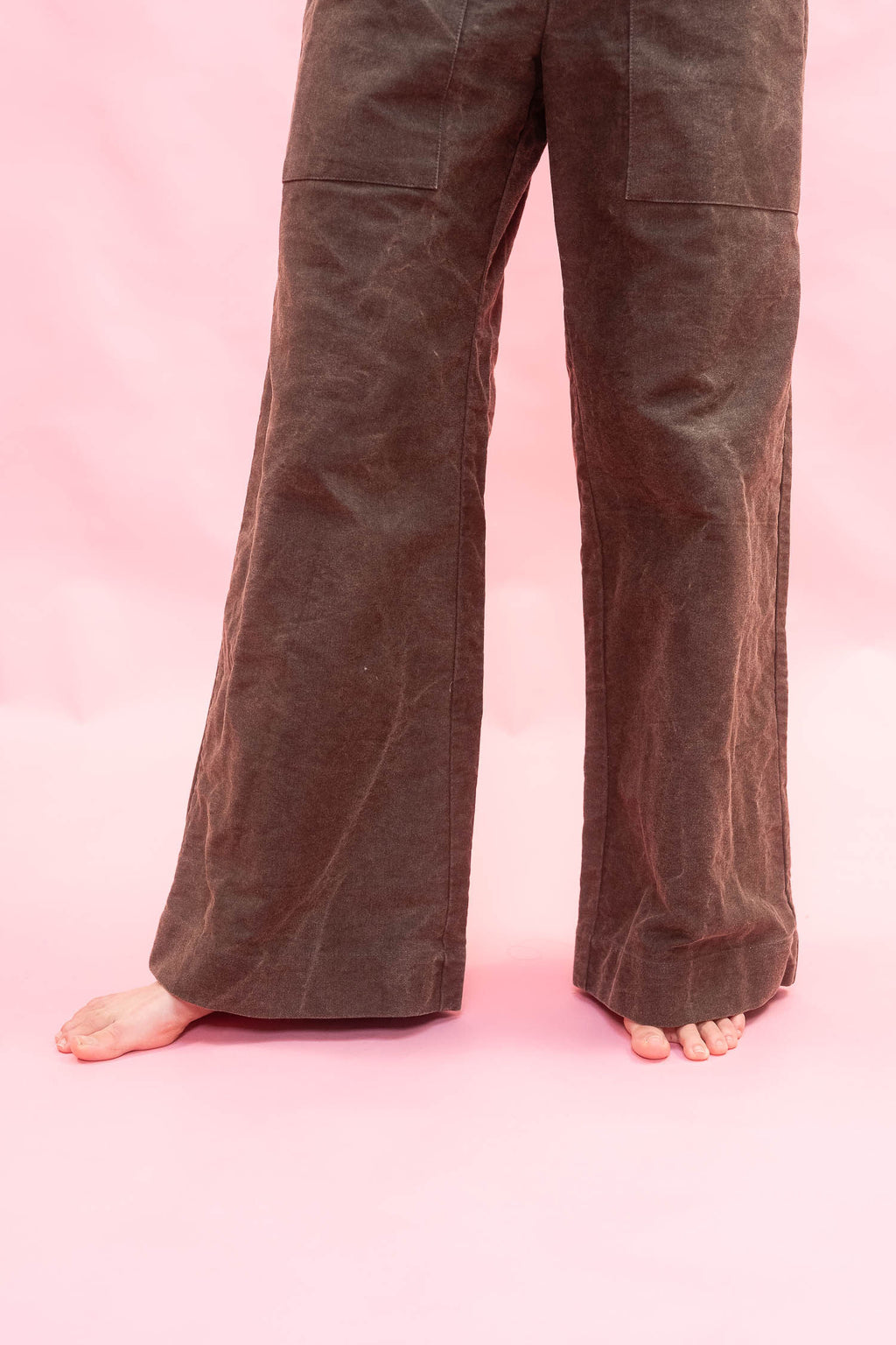 Brown wide-leg cotton
 pants shown on a model from the waist down, standing barefoot against a solid pink background.