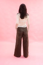 A woman viewed from behind wearing a white long-sleeve top and dark brown wide-leg cotton pants, standing against a solid pink studio backdrop.