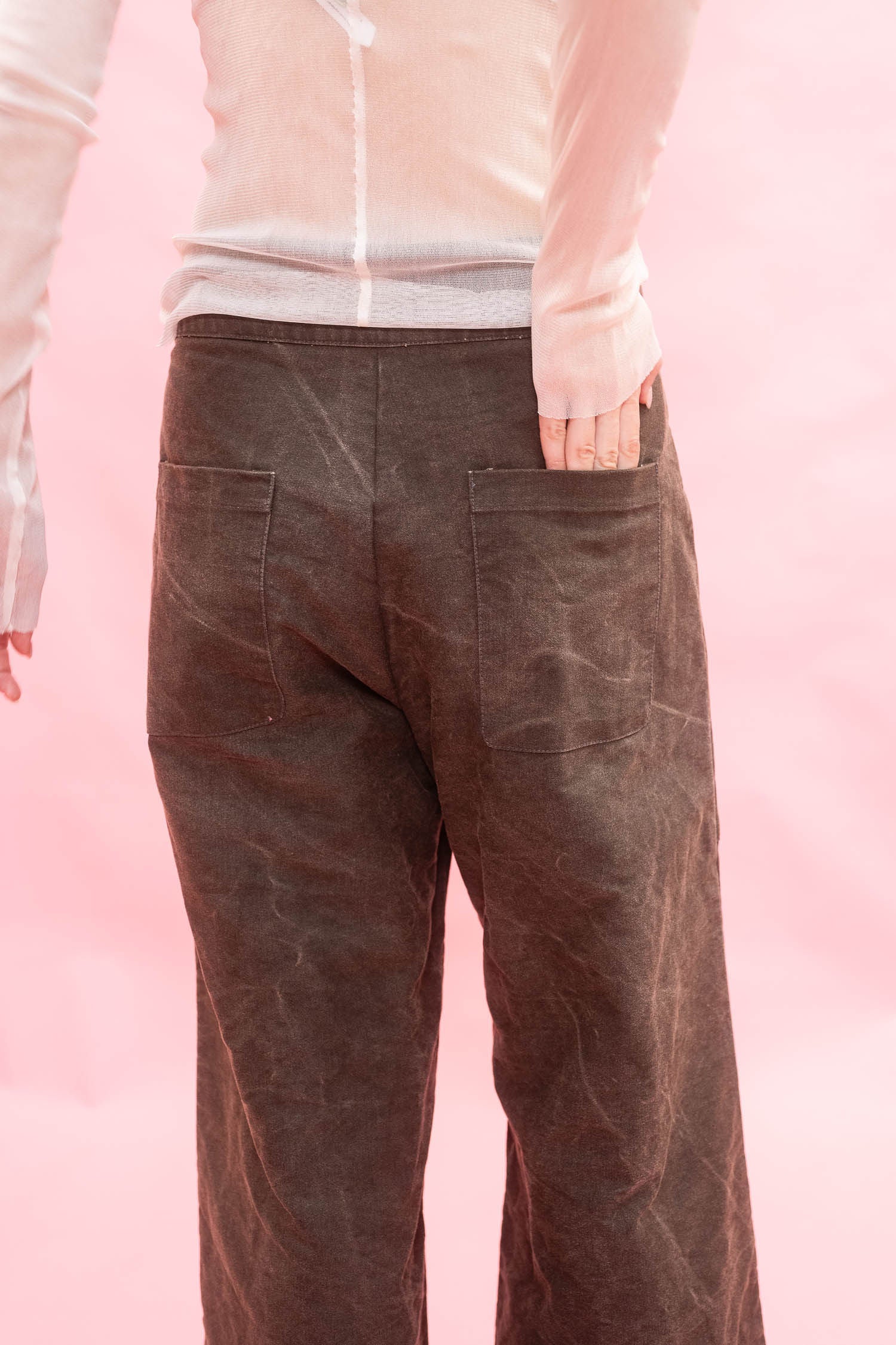 A person shown from the back wearing dark brown wide-leg pants with a textured, waxed finish and a light pink long-sleeve top, set against a solid pink background.