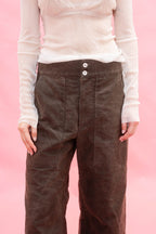 A person wearing dark brown wide-leg cotton pants with a white long-sleeve top, shown from the chest down against a solid pink background.